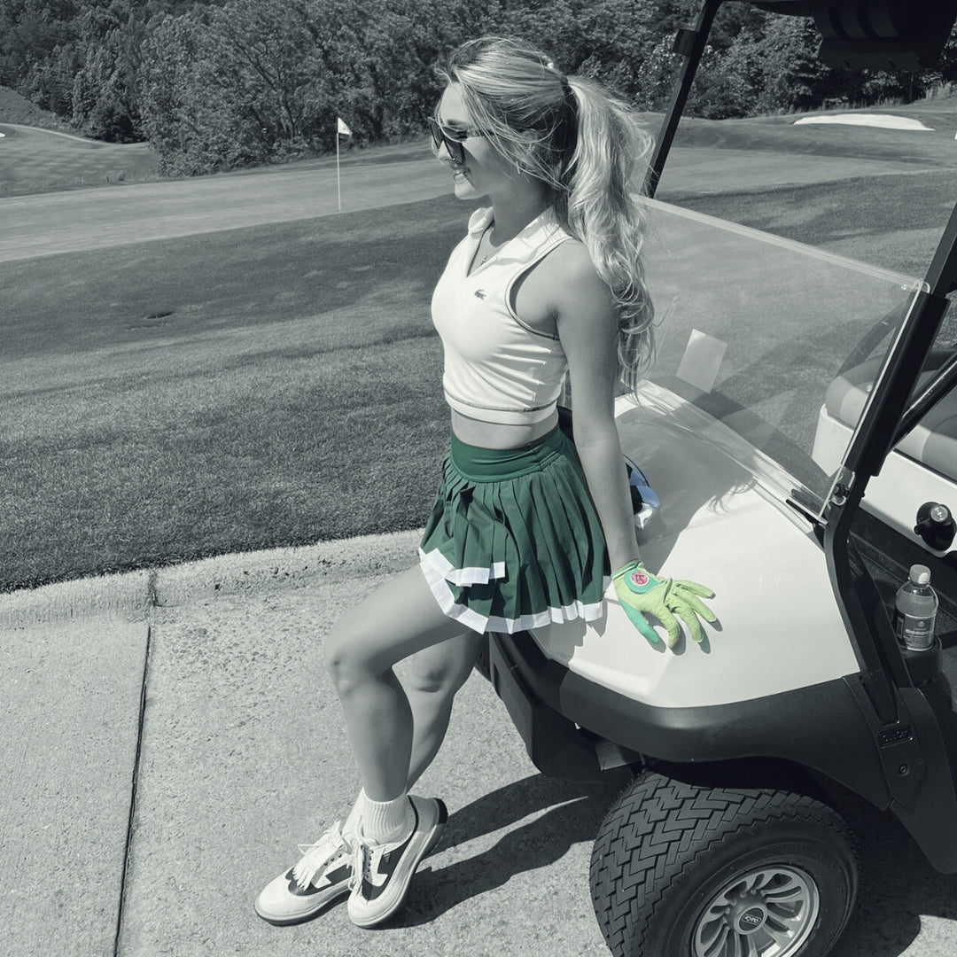 Woman in green skirt leaning on golf cart with her VivanTee green golf glove with ball marker in a monochromatic background.