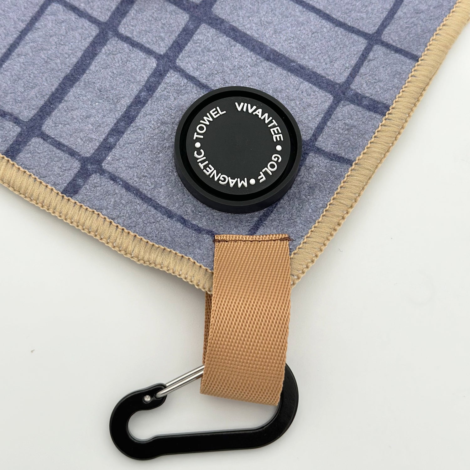 Close-up image of a luxury golf towel featuring a soft gray fabric with a subtle blue grid pattern. The towel is accented by a tan loop with a black carabiner for easy attachment. A sleek, round magnetic marker is attached, displaying the text "VIVANTEE GOLF • MAGNETIC • TOWEL" in a white, elegant font, reinforcing the premium quality and modern design of the product. The overall aesthetic is refined and functional, embodying a sophisticated touch for the discerning golfer.