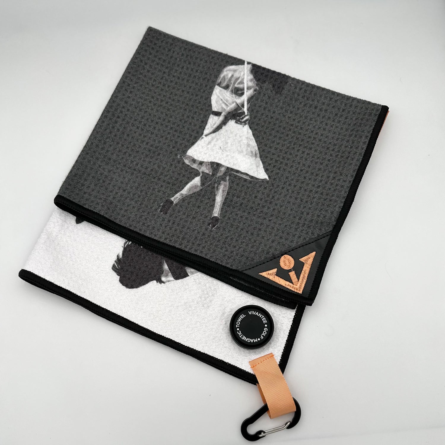 An elegant golf towel folded to showcase its unique design, featuring a grayscale image of a female golfer mid-swing, printed on a textured black fabric. The towel's corner is adorned with a refined geometric logo in a soft tan color, adding a touch of luxury. A tan loop with a black carabiner provides practical functionality, while a sleek magnetic marker labeled "VIVANTEE GOLF • MAGNETIC • TOWEL" enhances the premium aesthetic. The combination of monochrome art and modern details conveys a sense of timele