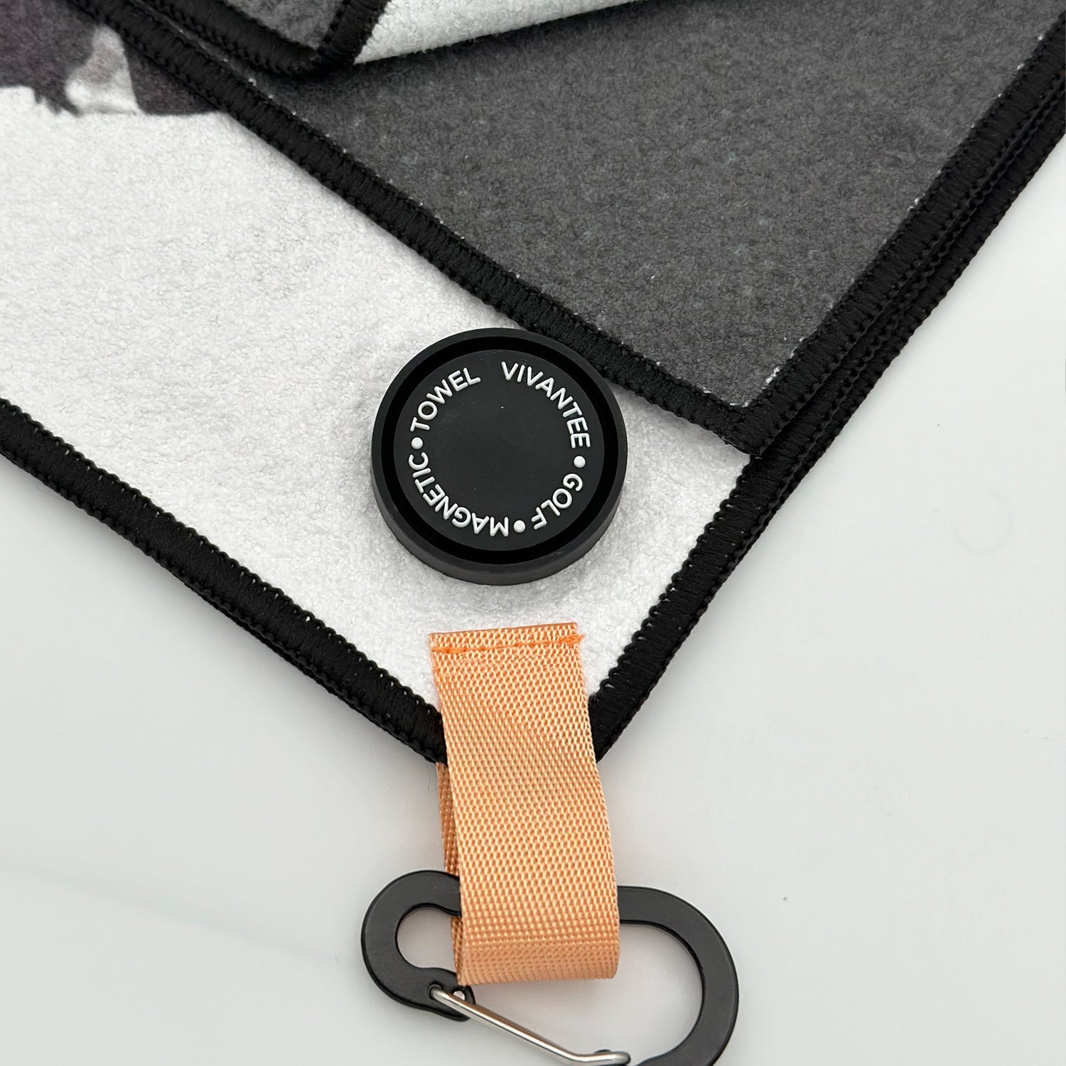 Close-up image of a luxury magnetic golf towel featuring a white and black color theme. The towel is accented by an orange loop with a black carabiner for easy attachment. A sleek, round magnetic marker is attached, displaying the text "VIVANTEE GOLF • MAGNETIC • TOWEL"