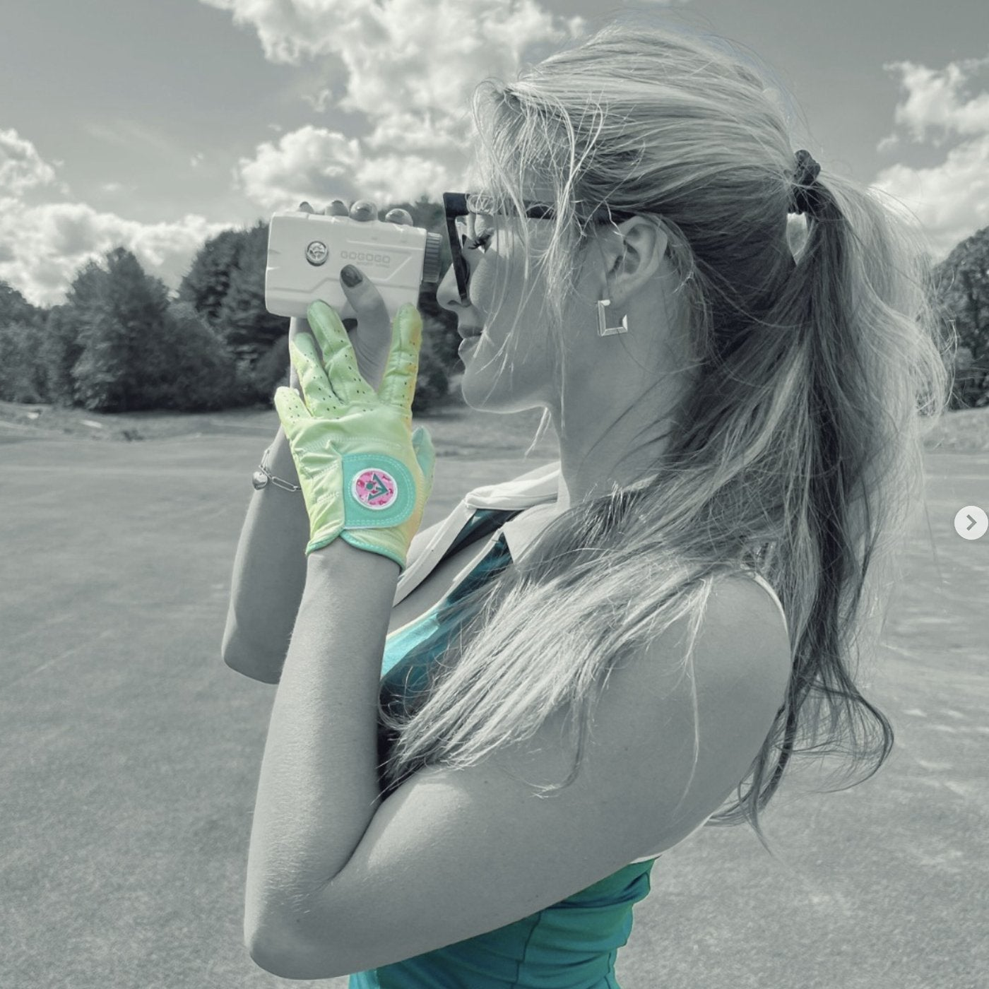 Woman holding a range finder while wearing a unique golf glove with ball marker in pink with a green dress.