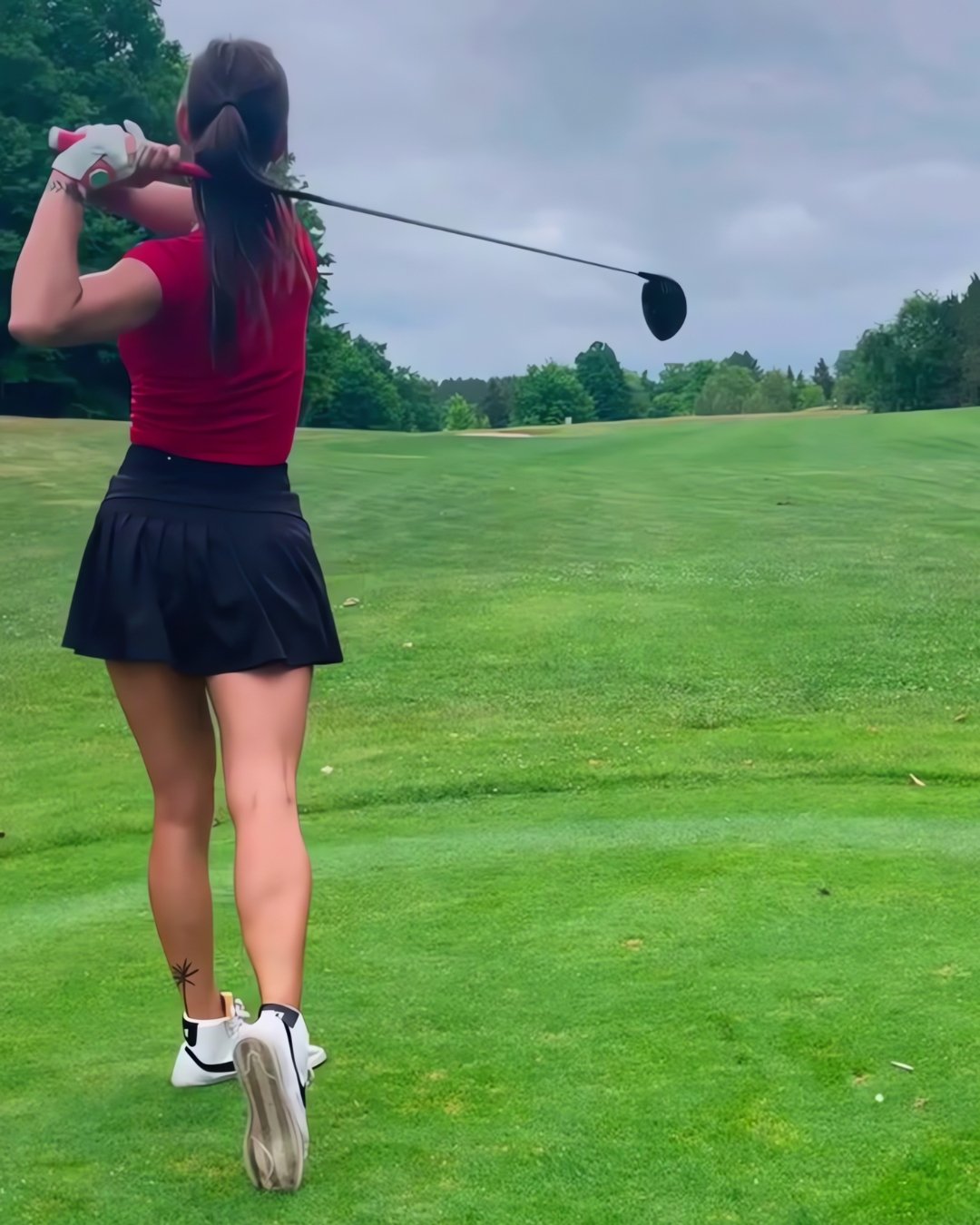 Stylish female golfer mid golf swing in red shirt and black skirt with a designer pink golf glove with magnetic ball marker.