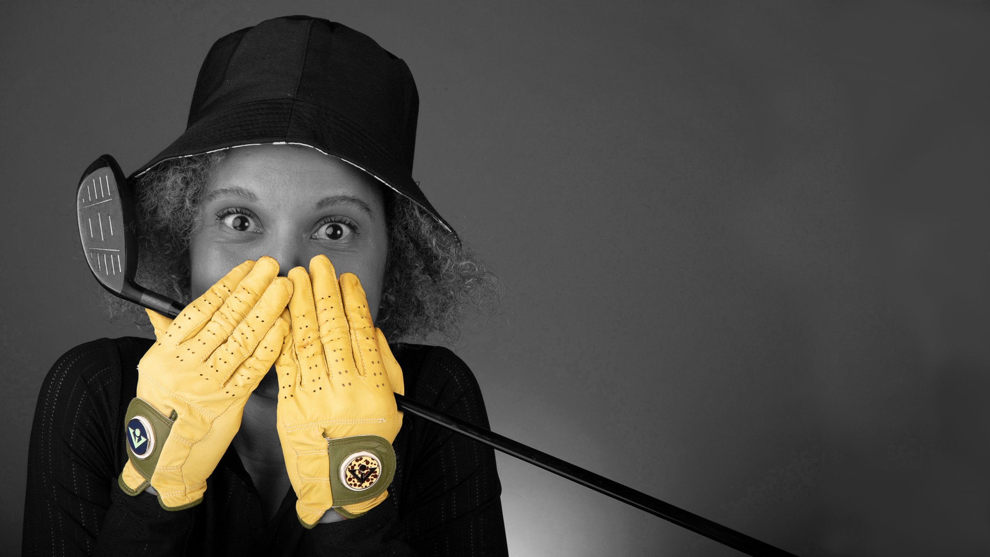 Woman wearing bright yellow unique golf gloves with magnetic ball markers, posing with a golf club and a bucket hat in a monochromatic background.